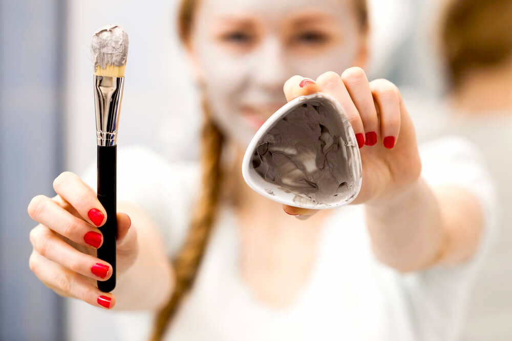 Young girl holding up clay mask and a face brush
