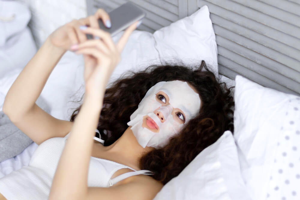 Woman with sheet mask, taking a selfie in bed 